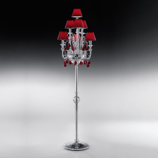 The Traditional Floor Lamp With Red Crystal Drops is an elegant silver fixture standing tall with multiple red lampshades and decorative red crystals. This classic, luxurious floor lamp features intricate detailing and rests on a shiny, broad metallic base, all showcased against a gradient black and white background.