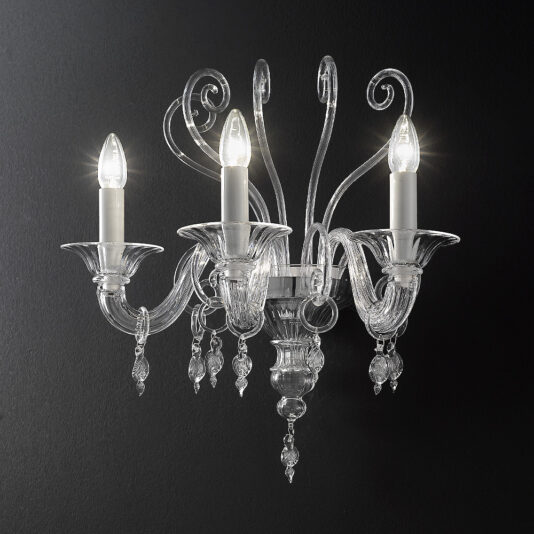 The Traditional Three-Armed Clear Glass Wall Light, mounted on a dark wall, showcases intricate scrollwork and hanging crystal ornaments. Each of its three arms holds a lit candle-shaped light bulb, casting a bright glow against the dark background.