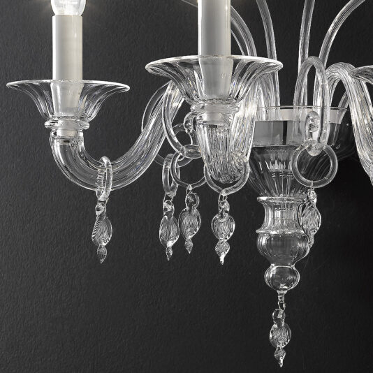 A close-up image of the Traditional Three-Armed Clear Glass Wall Light showcases its ornate design, with multiple curved arms that evoke elegance. Each arm supports candle-shaped light bulbs and decorative hanging glass pieces, beautifully illuminated against a dark background.