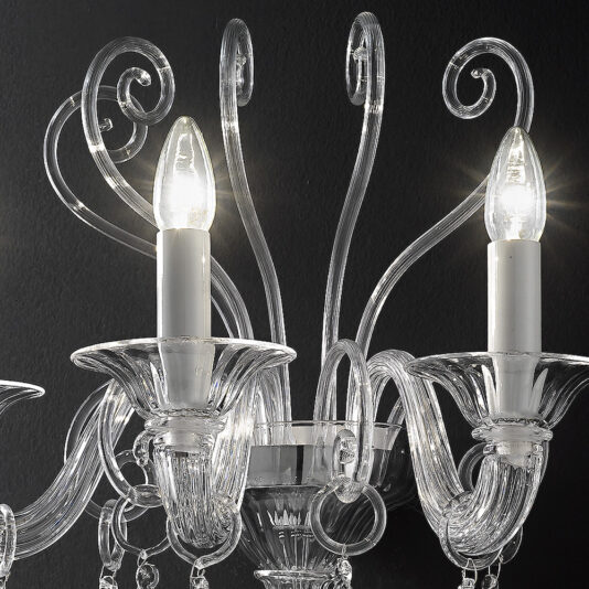 An elegant crystal chandelier against a black background features intricate, curved glass arms, complemented by the Traditional Three-Armed Clear Glass Wall Light. Adorned with clear candle-like holders that contain bulbs emitting a soft white light, the delicate design creates an ornate and luxurious appearance.