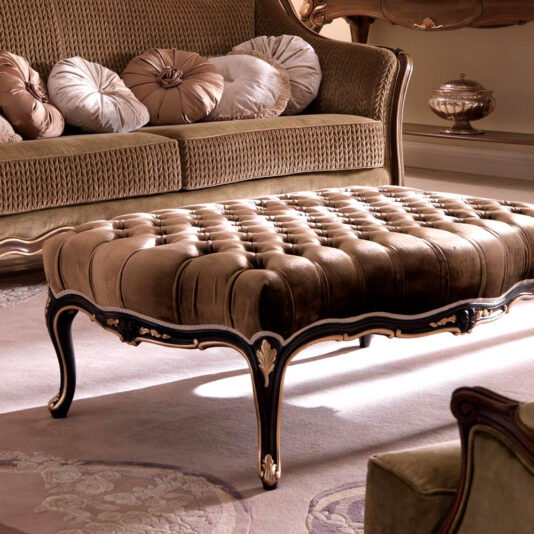 A luxurious living room featuring a Venetian Style Button Upholstered Coffee Table with ornate wooden legs, set on a plush rug. In the background, there's an elegant sofa adorned with multiple round, decorative pillows. The room exudes a classic, elegant ambiance.