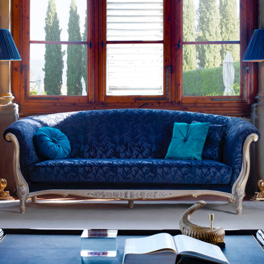 A luxurious room showcases the exquisite Venetian Style Classic Sofa in deep blue, featuring sophisticated carvings on its frame. The sofa is placed near a large window with a scenic view of tall trees and lush greenery. Adorned with teal and blue accent pillows, the room also boasts stylish lighting and decor elements to complete the elegant setting.
