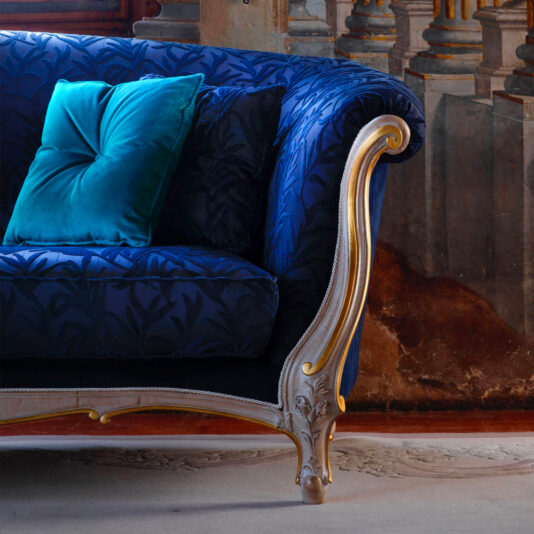 Venetian Style Classic Sofa The Venetian Style Classic Sofa exudes luxury with its wooden frame and intricate gold accents. It is upholstered in deep blue fabric featuring a leafy pattern and comes adorned with two teal pillows. The background showcases a mural with architectural elements in a rustic color palette, perfectly complementing the sofa's opulent design.