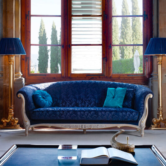 Venetian Style Classic Sofa A luxurious living room with a large window featuring a scenic view hosts the Venetian Style Classic Sofa, adorned with blue and turquoise pillows. The sofa is flanked by ornate gold floor lamps with blue shades. A coffee table holds an open book and decorative items, creating an inviting ambiance.