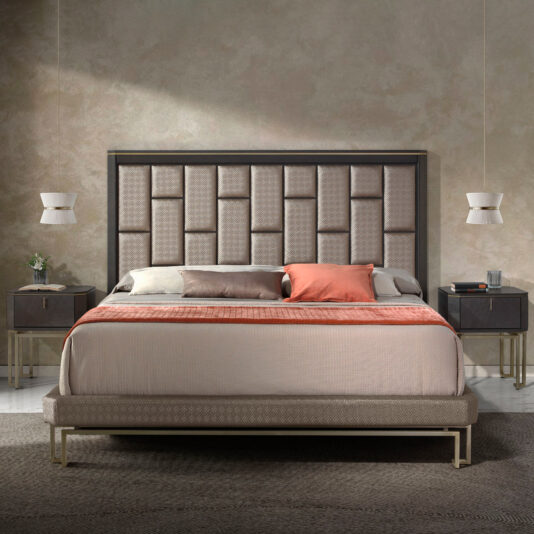 Modern Geometric Veneer Bedside Table A contemporary bedroom showcases a large bed with a textured, upholstered headboard, complemented by neutral bedding and an orange accent pillow. Flanking the bed are two Modern Geometric Veneer Bedside Tables, each illuminated by pendant lights hanging above. The floor is covered with a textured gray rug.