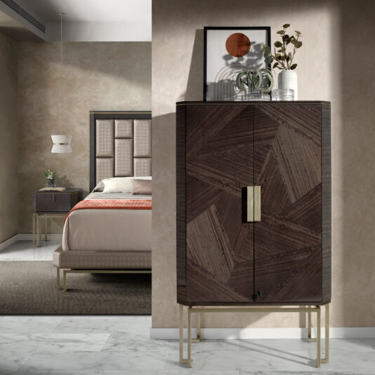 Modern Geometric Veneer Bedside Table A modern bedroom featuring a beige bed with a padded headboard, white bedding, and a brown throw blanket. Next to the bed is a small bedside table with a white lamp. In the foreground, there is a Modern Geometric Veneer Bedside Table adorned with decorative items on top.