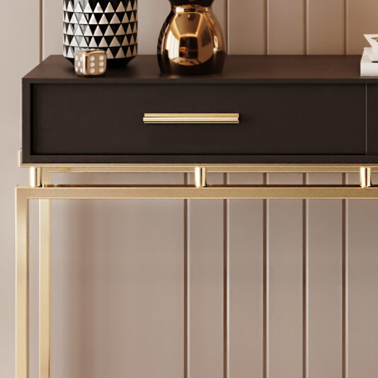 The Art Deco Inspired Console Table, showcasing a black drawer accented with a gold handle, stands against a beige paneled wall. The table features an elegant gold geometric base and is adorned with a black-and-white patterned vase, a small square object, and a reflective gold vase.
