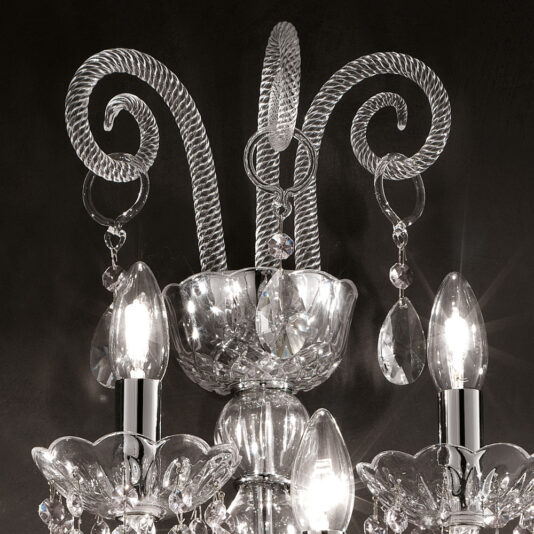 Classic Glass Wall Light With Crystal Drops A close-up view of the Classic Glass Wall Light With Crystal Drops against a dark background. This ornate fixture showcases intricate glasswork with elegant curving details, adorned with hanging crystal ornaments and three candle-like bulbs that emit a bright light.