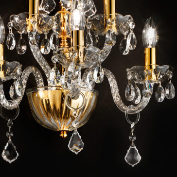 A close-up of an ornate chandelier featuring golden accents and clear, intricately cut glass components. The chandelier, evocative of the Classic Gold 5 Arm Wall Light With Crystal Drops, boasts multiple candle-like bulbs, crystal droplets, and a polished golden bowl set against a black background, casting a warm, elegant glow.