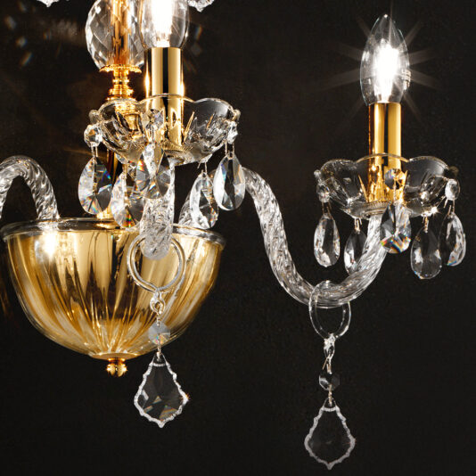 Classic Gold Wall Light With Crystal Drops A close-up of the Classic Gold Wall Light With Crystal Drops reveals its luxurious chandelier-like elegance, featuring gold accents and gleaming crystal decorations. The intricate designs are highlighted by a bright light bulb, casting a warm glow on the surrounding crystal droplets against a dark background.