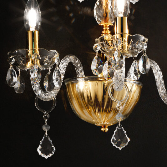 Classic Gold Wall Light With Crystal Drops A close-up of a Classic Gold Wall Light With Crystal Drops reveals its luxurious chandelier design featuring intricate golden and crystal elements. The multiple ornate arms each cradle a candle-style light bulb, exuding an air of timeless elegance. Elegant crystal pendants dangle from the fixture, capturing and reflecting light beautifully against the dark background.