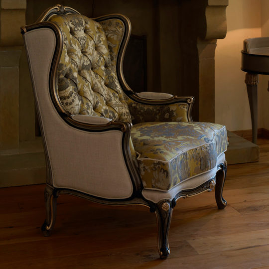 The Classic High Backed Venetian Style Armchair features intricate woodwork and elegant floral upholstery. It sits gracefully on a polished wooden floor, adjacent to a stone fireplace and a small side table, with warm lighting accentuating the cozy, vintage ambiance of the room.