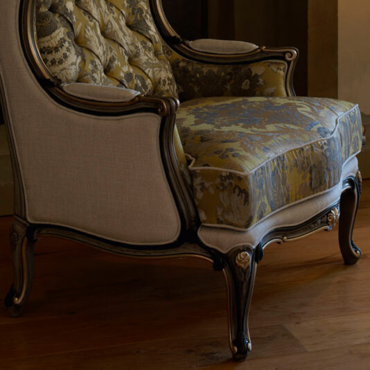 The Classic High Backed Venetian Style Armchair is an ornate, vintage piece with a carved wooden frame. This elegant chair features a beige cushioned backrest and a floral patterned seat cushion in shades of gold and light blue, set against a wooden floor.