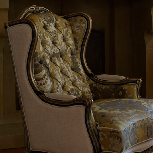 The Classic High Backed Venetian Style Armchair, adorned with intricate floral upholstery and supported by a dark wooden frame, boasts a tufted backrest and elegantly curved armrests. It’s ideally situated in a dimly lit room exuding a warm, rustic ambiance.