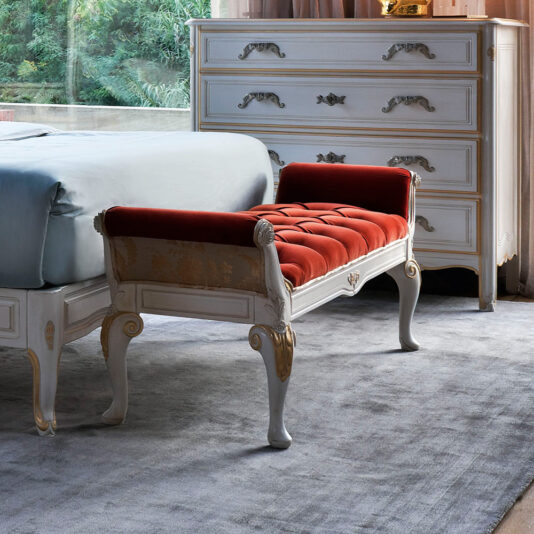 An elegant bedroom boasts a white wooden dresser adorned with intricate details and gold accents, a Classic Louis Style Button Upholstered Bench featuring rich red upholstery at the foot of the bed, and a view of lush greenery through the window. The bed is dressed in light blue bedding, and the floor is covered in gray carpet.