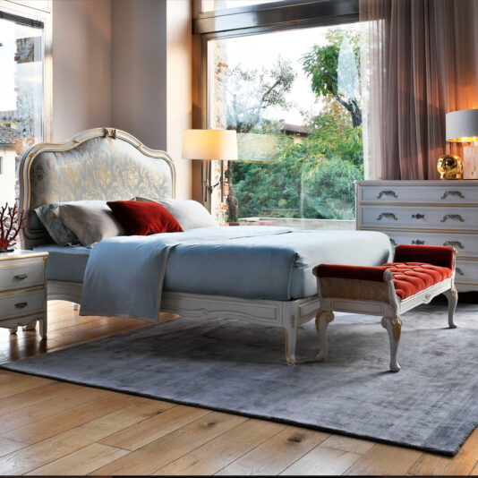 Elegant bedroom with large windows providing natural light. Features a vintage-style bed with a high, ornate headboard, blue bedding, and red accent pillows. A Classic Louis Style Button Upholstered Bench with a red cushion stands at the foot of the bed. The room has a chest of drawers and bedside tables.