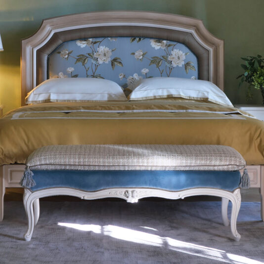 A neatly made bed with a floral-patterned upholstered headboard, flanked by two pillows. A **Classic Louis Style Upholstered Bench** with ornate legs sits at the foot of the bed. The room features a green wall and natural lighting, with a small plant on a side table.