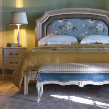 Classic Louis Style Upholstered Bench An elegant bedroom showcases a decorative bed adorned with a floral headboard and layered with mustard and white bedding. At the foot of the bed, the Classic Louis Style Upholstered Bench adds timeless charm. A vintage nightstand, paired with a lamp casting a warm glow, completes the classic decor on top of a plush carpet.