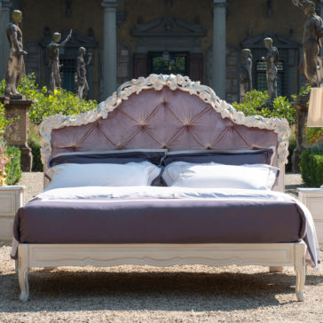An elegant outdoor scene showcases the Classic Style Button Upholstered Bed With Carvings, adorned with white and purple bedding. Positioned in a historic garden surrounded by weathered statues and lush greenery, this luxurious bed creates an exquisite and tranquil atmosphere.