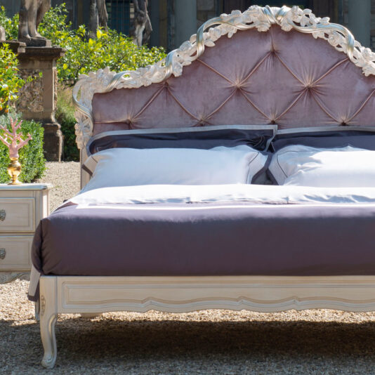 Classic Style Button Upholstered Bed With Carvings A luxurious Classic Style Button Upholstered Bed With Carvings, featuring a tufted headboard in plush light pink fabric, is elegantly set outdoors. The bed is adorned with crisp white sheets and a dark gray comforter. Beside it stands a small vintage bedside table accented in matching white and gold tones.