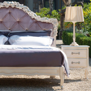 The Classic Style Button Upholstered Bed With Carvings, showcasing an ornate, tufted headboard and plush pillows, is elegantly positioned outdoors next to a white vintage-style nightstand with a lamp. The setting includes lush greenery and a stately stone building in the background, evoking the charm of an elegant garden or courtyard.
