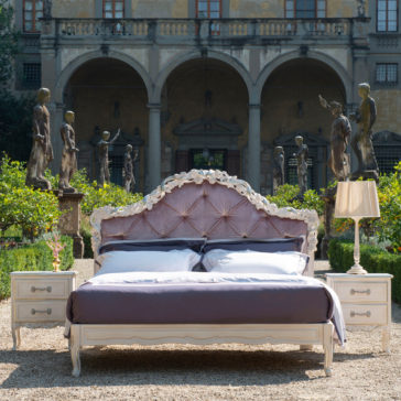 A luxurious Classic Style Button Upholstered Bed With Carvings and white linens is placed outdoors in front of a grand building with arched windows and statues. Two bedside tables with lamps are positioned on either side, and lush greenery surrounds the scene.