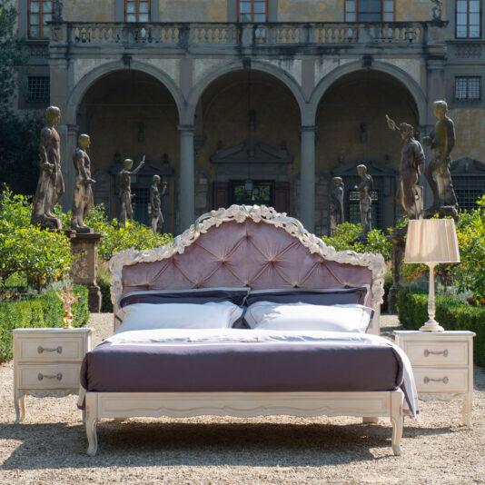 Classic Style Button Upholstered Bed With Carvings A luxurious Classic Style Button Upholstered Bed With Carvings and white linens is placed outdoors in front of a grand building with arched windows and statues. Two bedside tables with lamps are positioned on either side, and lush greenery surrounds the scene.