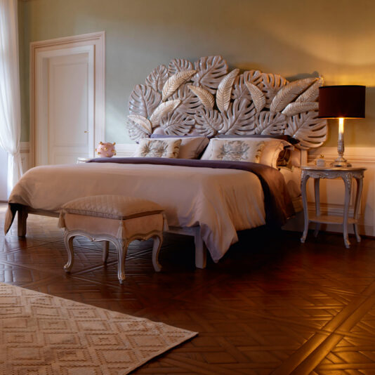 An elegant bedroom features a bed with a decorative headboard resembling large tropical leaves, flanked by a stylish nightstand with a black lampshade. The room, embodying classic style, has wooden flooring, a light-colored area rug, and the Classic Style Louis Small Bench Footstool at the foot of the bed.