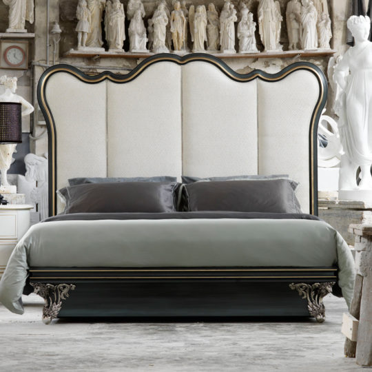 An ornate bedroom featuring the Classic Venetian Style Bed With Tall Headboard, showcasing a luxurious, tall, white upholstered headboard accented by black edging and intricate carvings on the legs. The bed is dressed in gray and black bedding, set against a backdrop of white classical statues and a clock on the left wall.