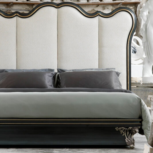 Classic Venetian Style Bed With Tall Headboard Close-up of a luxurious Classic Venetian Style Bed With Tall Headboard. The headboard showcases a large padded section with light patterned fabric upholstery, framed by dark wood with ornate edges. The bed is adorned with grey and black pillows and set against a stylishly rustic background.