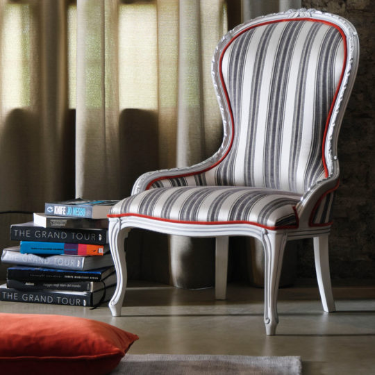 A Classic Venetian Style Occasional Chair featuring black, white, and red vertical stripes and elegant white wooden legs is positioned beside a stack of large hardcover books near a window shrouded in curtains. A glimpse of an orange cushion can be seen in the foreground.