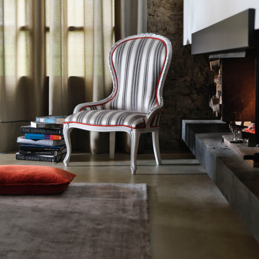 A cozy room features a Classic Venetian Style Occasional Chair positioned next to a window adorned with curtains. A stack of books rests beside the chair, and a red cushion is placed on the floor. To the right, a modern fireplace with glowing firewood enhances the comfortable ambiance.