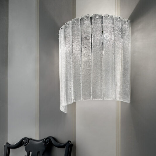 Demi Glass Wall Light A modern Demi Glass Wall Light featuring vertical, textured glass panels graces a soft grey wall. Just below and slightly to the left is a chair with a black, intricately designed backrest, creating an elegant and contemporary interior design scene.