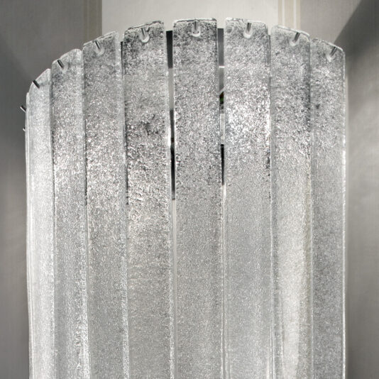 Demi Glass Wall Light A detailed close-up showcases several overlapping Demi Glass Wall Light panels, aligned vertically with a textured, frosted surface. These panels are suspended in a semi-circular formation, allowing light to pass through and generate a shimmering effect.