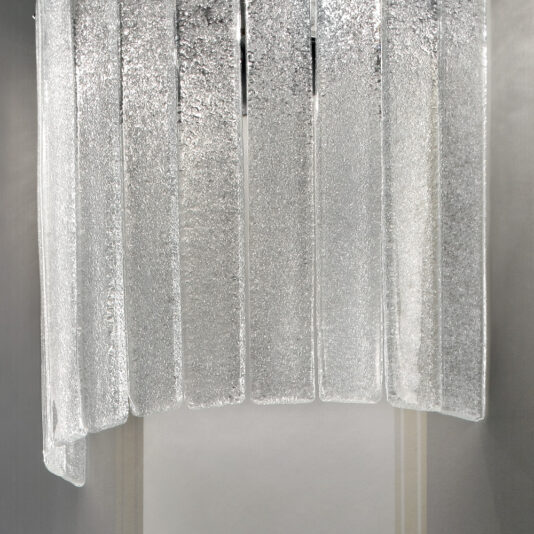 Demi Glass Wall Light Close-up image of a textured, vertical glass panel arrangement. Each panel is slightly curved and has a frosted, crystalline appearance, reminiscent of the Demi Glass Wall Light. This design creates a shimmering effect as the panels are arranged side by side against the neutral-colored wall.