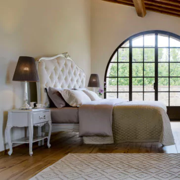 Elegant Classic Style Button Upholstered Bed A cozy bedroom featuring a large arched window, a king-sized Elegant Classic Style Button Upholstered Bed with an elegant tufted headboard, and a soft, neutral-colored comforter set. A bedside table with a lamp sits next to the bed, and an elegant area rug covers a portion of the wooden floor.