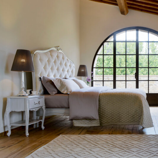 A cozy bedroom featuring a large arched window, a king-sized Elegant Classic Style Button Upholstered Bed with an elegant tufted headboard, and a soft, neutral-colored comforter set. A bedside table with a lamp sits next to the bed, and an elegant area rug covers a portion of the wooden floor.
