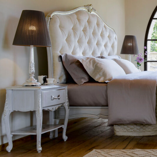 Elegant Classic Style Button Upholstered Bed A luxurious bedroom featuring the Elegant Classic Style Button Upholstered Bed adorned with tan and white bedding. Two ornate side tables with decorative lamps flank the bed, exuding an elegant classic style. The room has a wooden floor, beige walls, and a large arched window with an outside view partially visible.