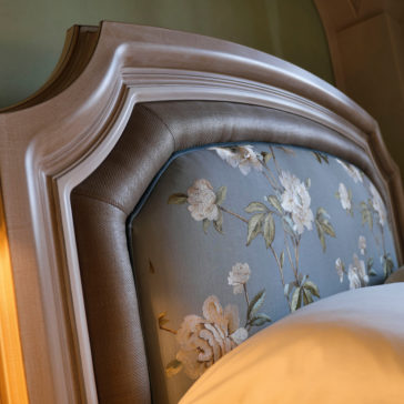 A close-up of the Elegant Classic Style Upholstered Bed reveals its sophisticated wooden headboard, featuring a luxurious upholstered panel with a delicate floral pattern. The design showcases white flowers and green leaves set against a light blue background. Intricate carvings and a polished finish adorn the headboard, which is partially covered by a white bedsheet.