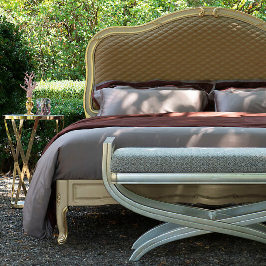 The Elegant Ornate Classic Style Bed features a quilted headboard and plush brown and gray bedding. A gold-accented side table with decor item completes the look, all amidst lush greenery.