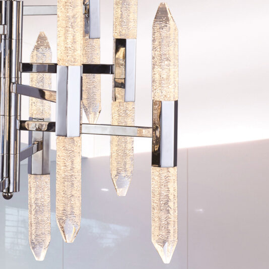 Exclusive Chandelier With Vertical Crystals The Exclusive Chandelier With Vertical Crystals features elongated, translucent crystal rods and silver metal accents. The textured crystals are illuminated, casting a warm glow against a minimalist white background.