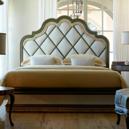 A luxurious bedroom featuring the **Exclusive Bed With Tassel Design Detail**, complete with an intricately quilted headboard, patterned pillows, and a golden bedspread. The room is adorned with elegant furniture, including a round side table and a chair. Large windows and white curtains create a bright atmosphere.