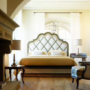 A luxurious bedroom featuring the Exclusive Bed With Tassel Design Detail, complete with a tufted, arched headboard adorned with decorative pillows. The room includes elegant side tables with lamps, an upholstered bench at the foot of the bed, and a fireplace. Natural light streams in through a large window with white curtains.