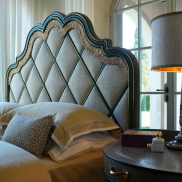 Elegant bedroom scene featuring the Exclusive Bed With Tassel Design Detail, boasting a large, ornate headboard with a quilted pattern. Beige and yellow bedding with decorative pillows adds charm. A bedside table with a lamp, small jar, and vase sits by an arched window with sheer curtains, letting in natural light.