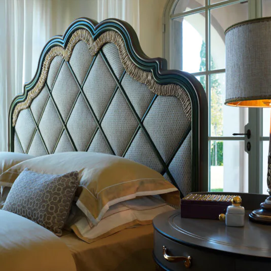Elegant bedroom scene featuring the Exclusive Bed With Tassel Design Detail, boasting a large, ornate headboard with a quilted pattern. Beige and yellow bedding with decorative pillows adds charm. A bedside table with a lamp, small jar, and vase sits by an arched window with sheer curtains, letting in natural light.