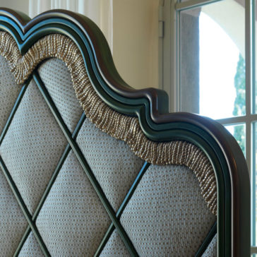 Close-up of the Exclusive Bed With Tassel Design Detail, featuring an ornate upholstered headboard with a wavy wooden frame and intricate gold accents. The fabric displays a striking diamond pattern and tassel designs. Sunlight streams through a window in the background, enhancing the bed's luxurious appearance.