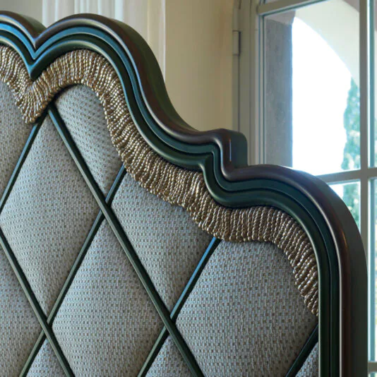 Close-up of the Exclusive Bed With Tassel Design Detail, featuring an ornate upholstered headboard with a wavy wooden frame and intricate gold accents. The fabric displays a striking diamond pattern and tassel designs. Sunlight streams through a window in the background, enhancing the bed's luxurious appearance.