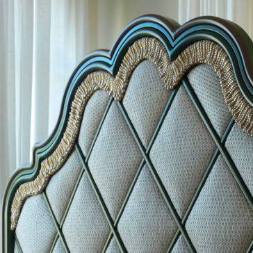 Close-up of the Exclusive Bed With Tassel Design Detail, showcasing a tufted, diamond-patterned fabric headboard. The headboard features a curved top edge adorned with decorative gold or metallic rope trim and tassel accents. White curtains hang in the background, adding a soft element to the scene.