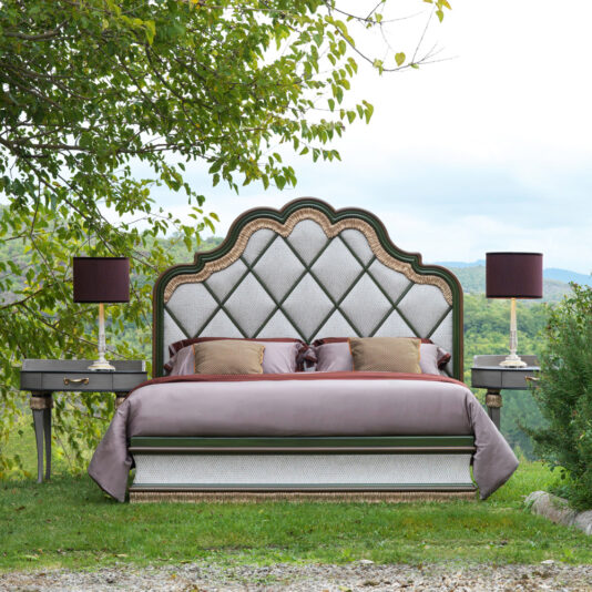 A luxurious bed outdoors on grass, surrounded by greenery, features an ornate quilted headboard and is flanked by Exclusive Bedside Tables with tassel design details and matching lamps. The backdrop includes trees and a hilly landscape.