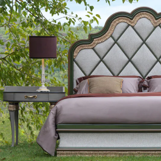 An exclusive outdoor bedroom setup showcases the Exclusive Bed With Tassel Design Detail, featuring a tufted headboard and adorned with pink and gray bedding. Beside the bed is an elegant gray nightstand with a drawer, topped by a purple lamp. The serene background reveals lush greenery and natural landscapes, enhancing the luxurious ambiance.